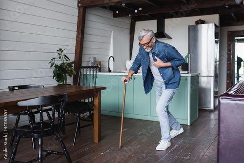 Sick senior man with crutch holding hand on chest at home