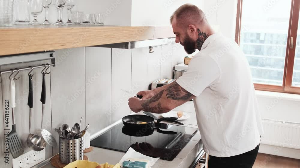 A side view of a muscular man is frying eggs standing in the kitchen at home A side view of a muscular man is frying eggs standing in the kitchen at home