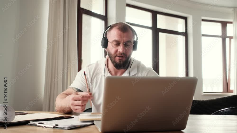 A serious man wearing headphones is talking with video connection sitting at home A serious man wearing headphones is talking with video connection sitting at home