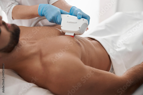 Master in blue gloves removes hair from chest of male client usunig modern equipment at photoepilation photo