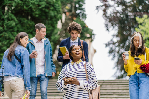 Positive brooding attractive african american female student thinking and looking upward standing on stairs while people, fellow students are walking around her.