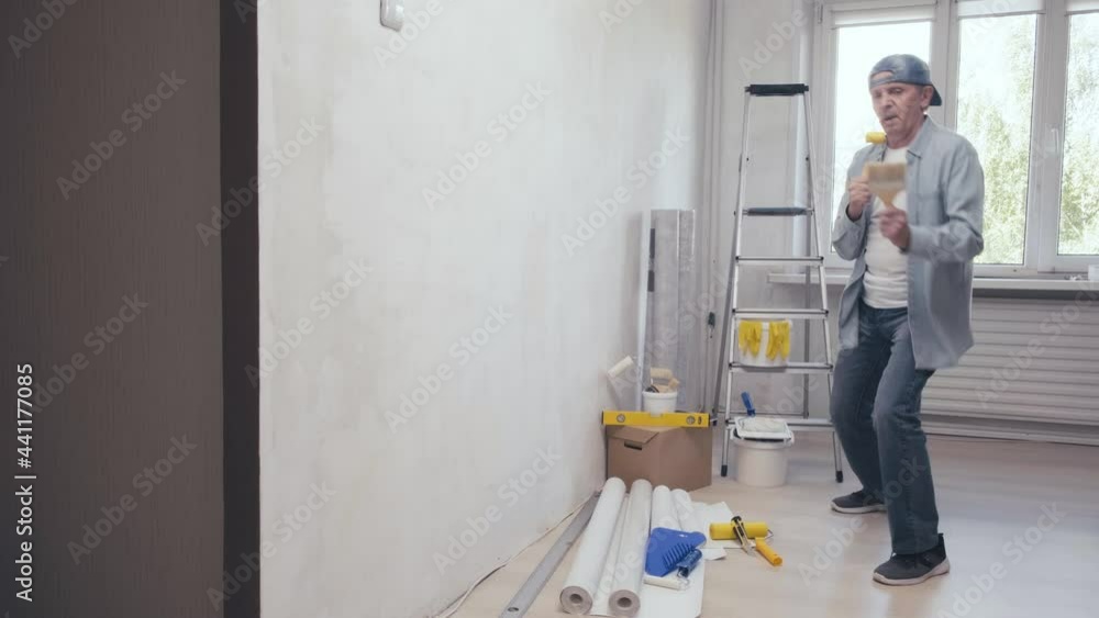 Positive man dancing and singing into a rubber roller having fun during home repair and painting. Construction site and renovation. House improvement and makeover concept. Room renovations at house Positive man dancing and singing into a rubber roller having fun during home repair and painting. Construction site and renovation. House improvement and makeover concept. Room renovations at house
