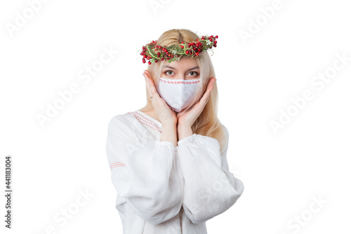 A blonde girl in a birch wreath and in a simple white linen dress with a protective mask on her face holds her hands at the face. Looks straight. Isolated on a white background