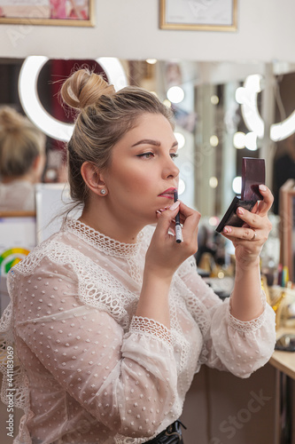 A beautiful blonde woman with hair taken away makes a make-up at the mirror in the dressing room, emphasizes the contour of the lips with a pencil