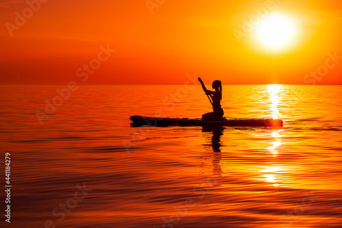 Girl on stand up paddle board at quiet sea with bright sunset or sunrise. Woman paddle on sup board in sea. © artifirsov