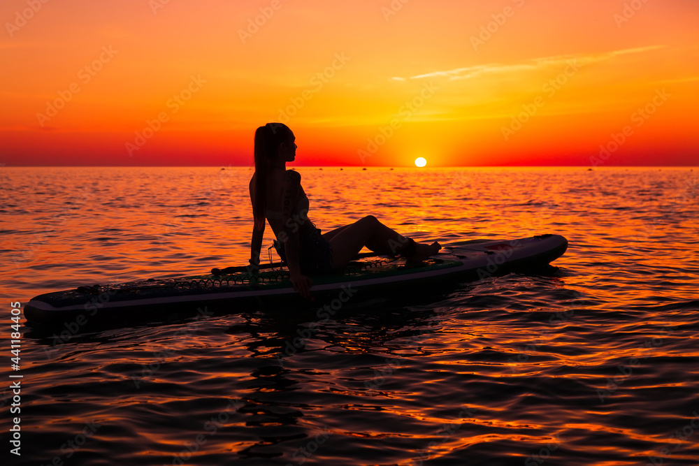 Girl on stand up paddle board at quiet sea with bright sunset or sunrise. Woman relaxing on sup board in sea.