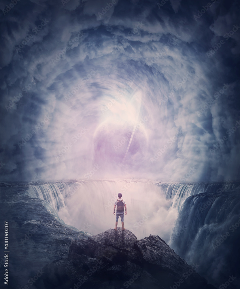 Naklejka premium Person stands on the edge of a cliff above a waterfall looking at a huge whirlwind in the clouds that creates a portal to another planet. Surreal and fantasy scene, magical world adventure concept