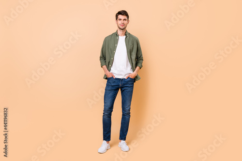 Photo of charming confident young guy dressed green shirt smiling walking arms pockets isolated beige color background