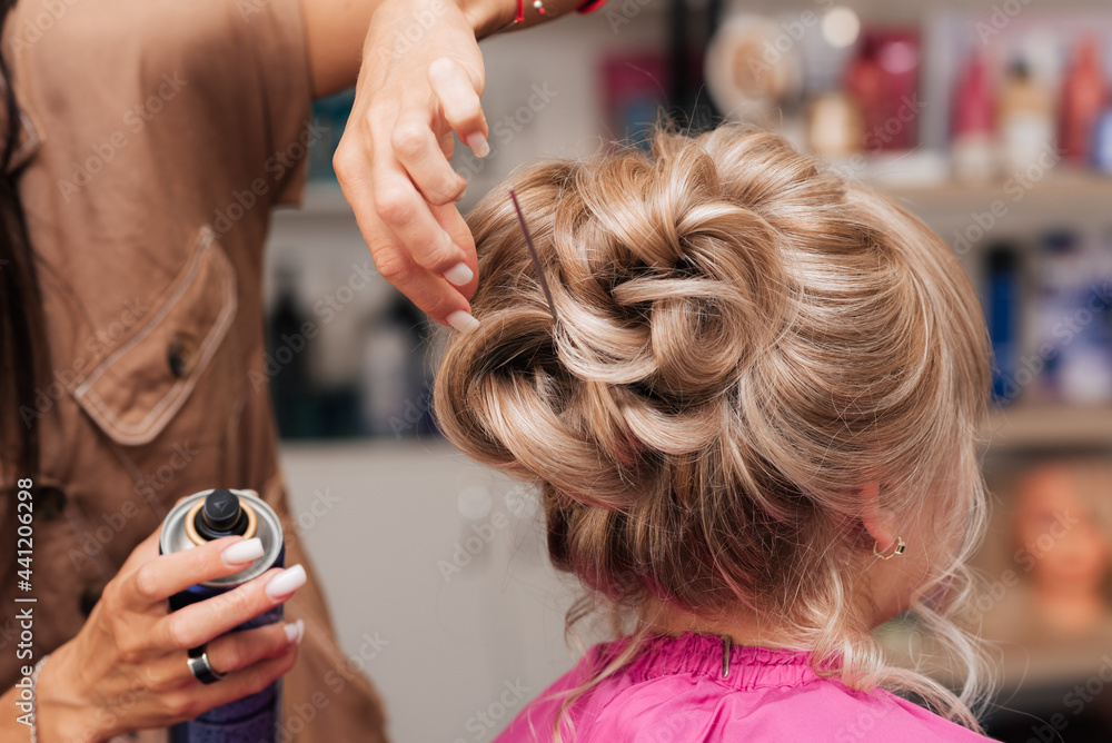 Fototapeta premium The girl-hairdresser makes the client a hairstyle for a celebration. The hairdresser fixes everything with hairspray