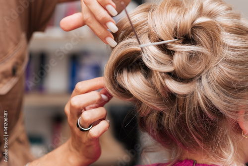 A girl-hairdresser makes a client's hairstyle for the celebration by pinning the strands with hairpins and hairpins.