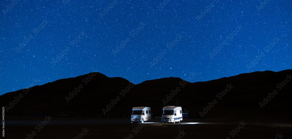 Campers parked in a caravan parking area sleeping on a starry night in the mountain. Summer tourism with RV in a blue night sky with stars. Best option for travel. Panorama motorhomes and camping car. - obrazy, fototapety, plakaty 