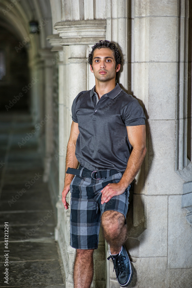 Dressing in a gray shirt, pattern shorts, a young strong handsome guy is standing by a old fashion wall, confidently looking forward.