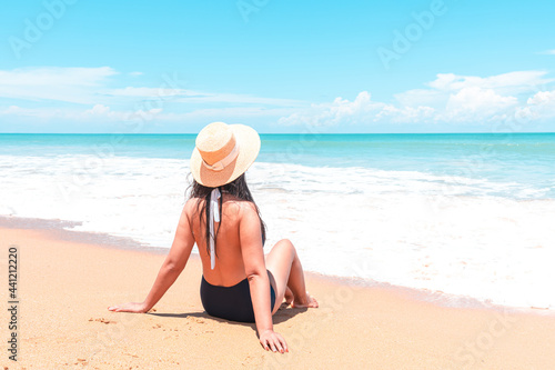 Back view of asian traveler woman in swimsuit and straw hat to sitting in the spray of the sea to looking view