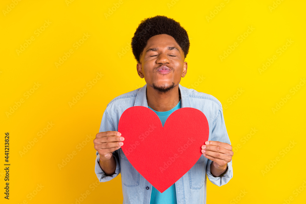 Photo of charming dreamy dark skin guy dressed denim shirt sending you kiss holding big red heart isolated yellow color background Photo of charming dreamy dark skin guy dressed denim shirt sending you kiss holding big red heart isolated yellow color background