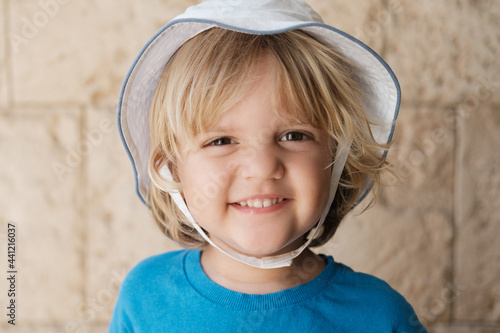 3 years old boy dressed in a blue t-shirt and white summer hat smiling at the camera on a sandstone background