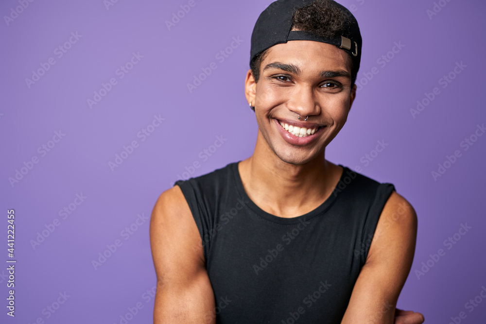 Portrait of smiling latino transgender man in black t-shirt and cap on purple background - obrazy, fototapety, plakaty 