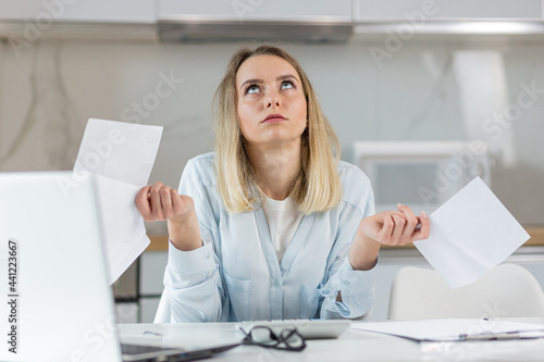 young female housewife sitting a table in house kitchen an incomprehensible deal with paperwork bills and loans. Woman is exhausted and frustrated with documents of enterprise or family budget at home