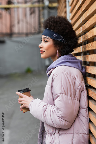 Side view of african american woman in bandana and jacket holding paper cup near wooden fence