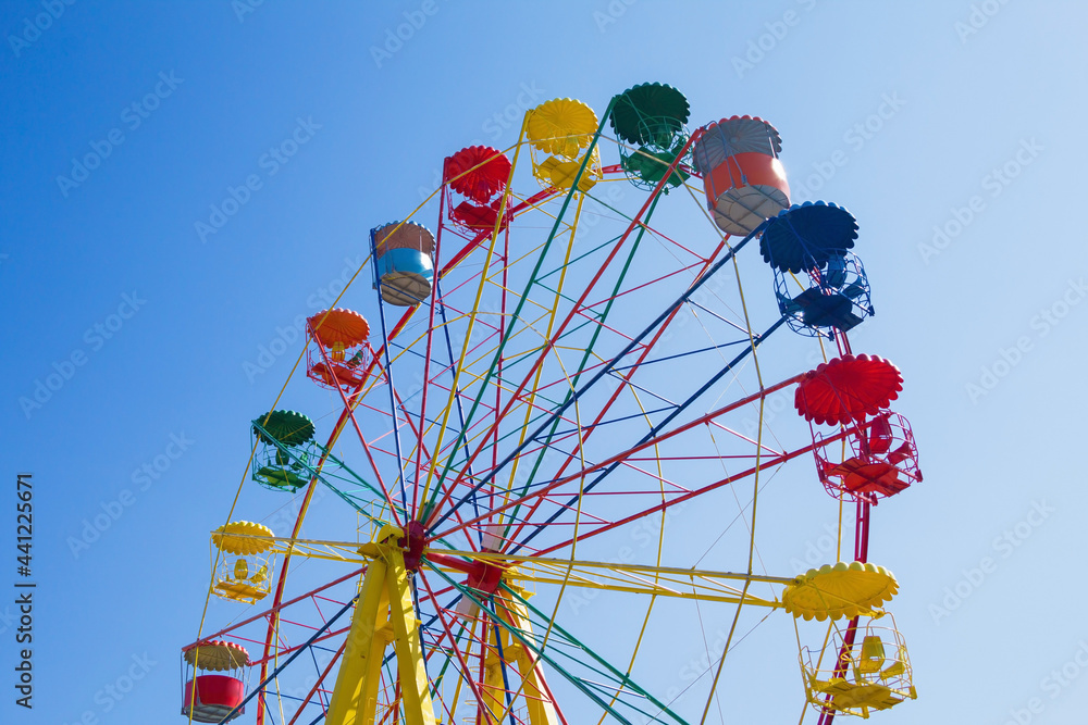 Bright multi-colored Ferris wheel against the blue sky. The wheel is shown with cabins. View from the ground below. Details of the attraction - red, yellow, green, blue. Copy space. 