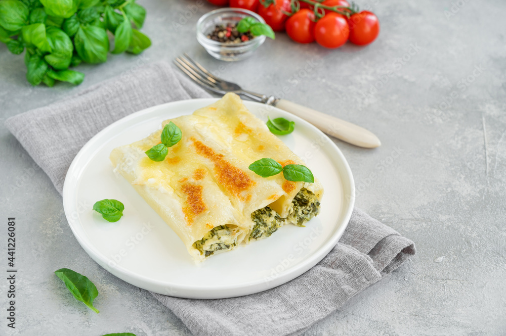 Traditional Italian cannelloni with ricotta and spinach in bechamel sauce with fresh basil leaves on a white plate on a gray concrete background. Vegetarian dish. Copy space.