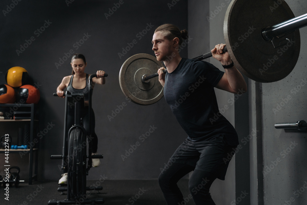 Couple have crossfit training at gym. Attractive fit woman is working out on exercise bike, while muscular man training with barbell. Functional and circuit training concept Couple have crossfit training at gym. Attractive fit woman is working out on exercise bike, while muscular man training with barbell. Functional and circuit training concept