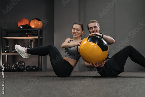 Healthy young couple doing abs exercises with medicine ball at dark gym. Couple sitting back to back on floor and doing workout by passing ball to each other. Sporty woman and man training together photo