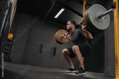 Muscular caucasian man lifting weights and doing back squat in gym. Crossfit athlete is holding a heavyweight barbell on the shoulder behind the neck while squatting. Fitness and work out
