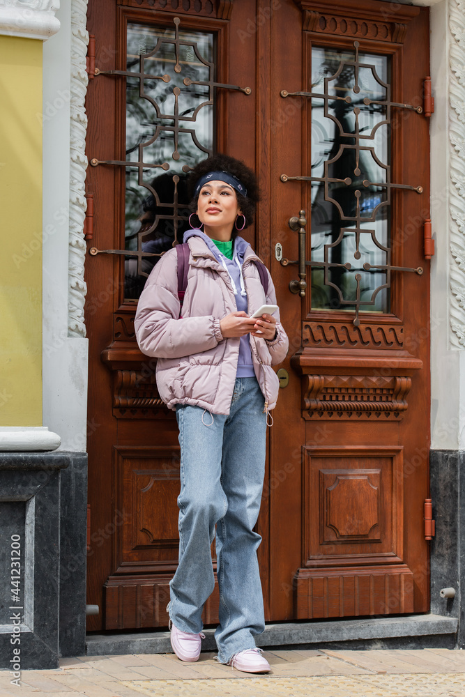 Fototapeta premium Young african american woman holding cellphone near door of building