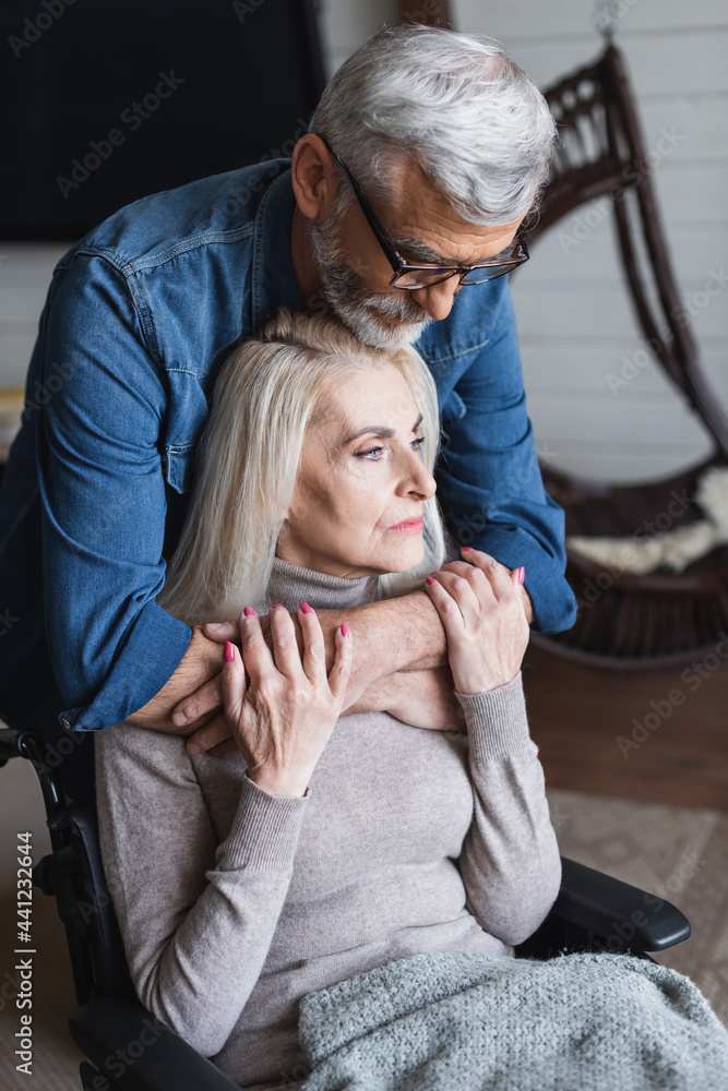 Fototapeta premium Careful man hugging wife in wheelchair at home