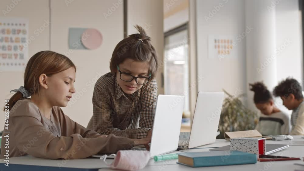 Medium slowmo of two teenage classmates chatting during break in multicultural school Girl typing something on laptop while talking to cheerful classmate Medium slowmo of two teenage classmates chatting during break in multicultural school Girl typing something on laptop while talking to cheerful classmate
