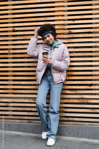 Smiling african american woman holding coffee to go near wooden fence