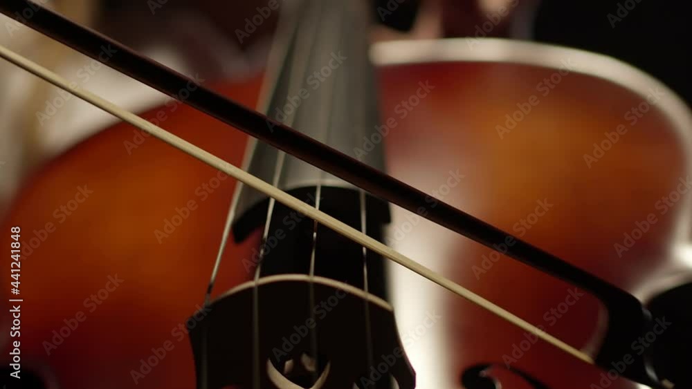 Bow plays slowly on cello strings, against a dark background. Light touches of bow to strings of wooden instrument. Concept art Bow plays slowly on cello strings, against a dark background. Light touches of bow to strings of wooden instrument. Concept art