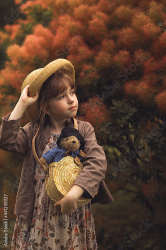 a girl in a hat with a toy teddy bear in her hands is standing in a beautiful summer garden