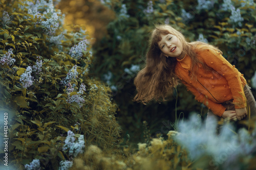 a girl with flowing hair looks out from behind the bushes of blooming lilacs and smiles
