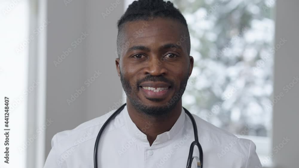 Close-up portrait of positive smiling African American male doctor posing indoors. Joyful handsome man in medical uniform looking at camera standing in hospital. Medicine and occupation Close-up portrait of positive smiling African American male doctor posing indoors. Joyful handsome man in medical uniform looking at camera standing in hospital. Medicine and occupation