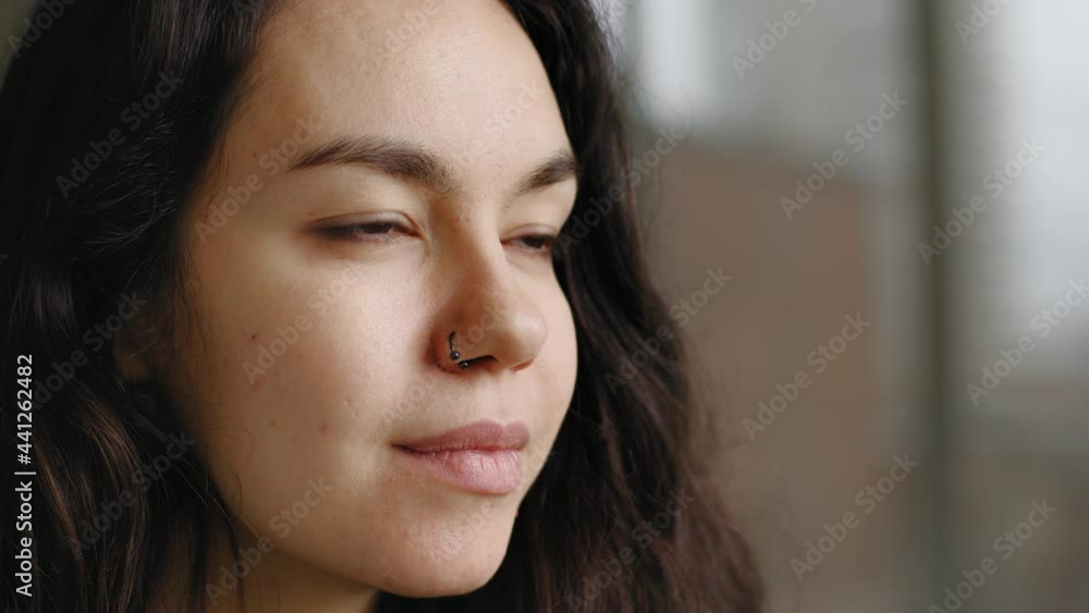 Extreme close-up female young face with perfect skin natural make-up dark eyes pierced nose earring posing looking away. Portrait caucasian millennial woman girl thinking contemplating in window Extreme close-up female young face with perfect skin natural make-up dark eyes pierced nose earring posing looking away. Portrait caucasian millennial woman girl thinking contemplating in window