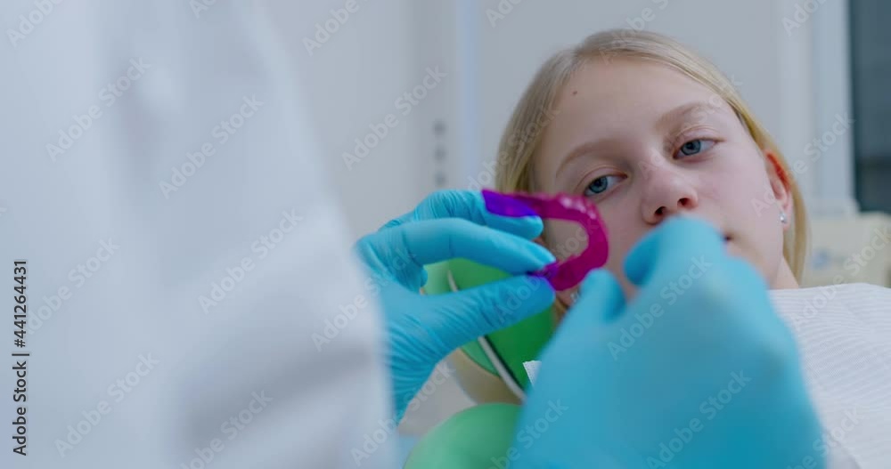 A little girl is sitting in the dentist's chair and listening to a dentist who is standing next to her and inserting a pink aligner into her lower jaw. A little girl is sitting in the dentist's chair and listening to a dentist who is standing next to her and inserting a pink aligner into her lower jaw.