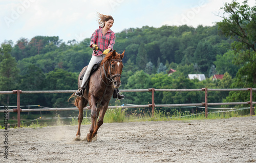 Young woman wearing shirt riding brown horse in sand paddock by wooden fence, hair moving in air because of speed