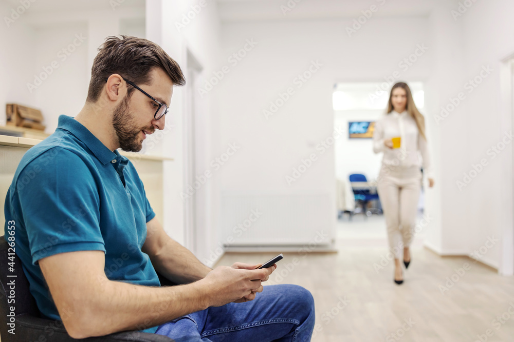 A man in waiting room. A man in casual clothes is sitting in the waiting room for the technical inspection and using a phone. In the background a blurred woman bringing a hot beverage A man in waiting room. A man in casual clothes is sitting in the waiting room for the technical inspection and using a phone. In the background a blurred woman bringing a hot beverage