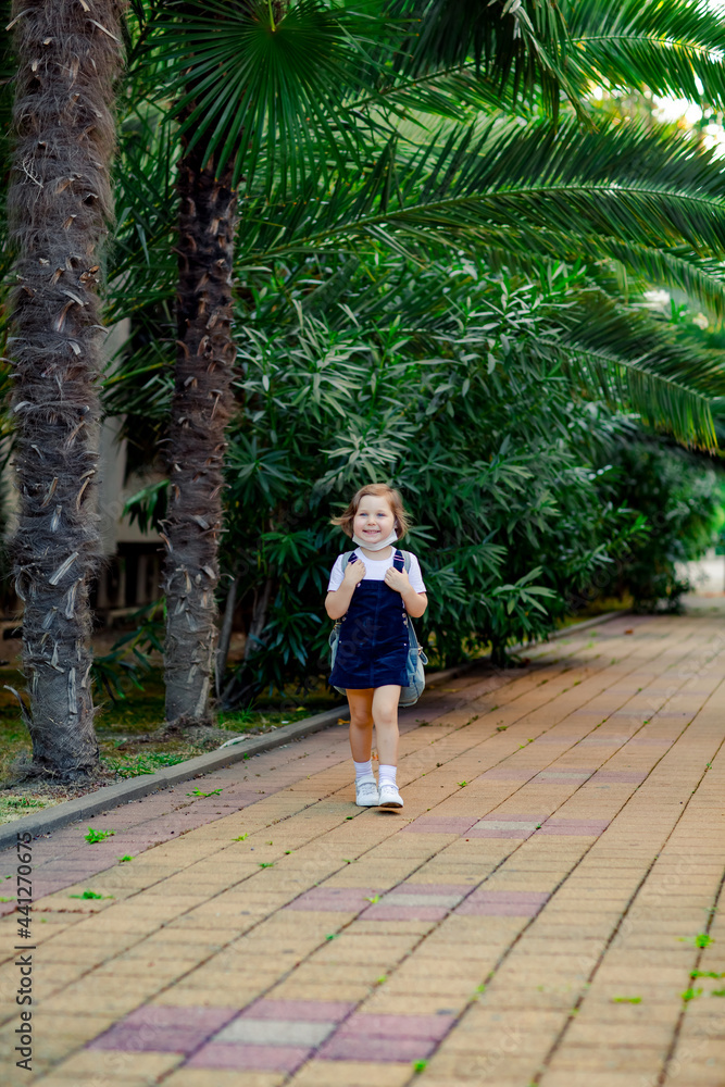 a little girl, a schoolgirl, goes to school on the street with a denim backpack, and in a medical mask