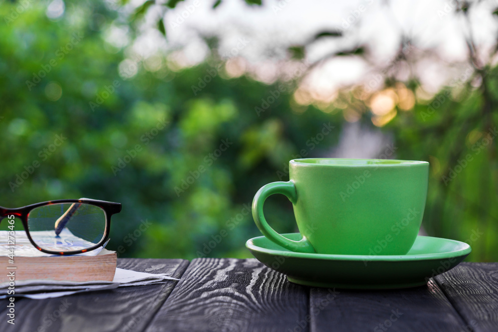 Green cup of hot coffee and glasses with a book and newspaper on a wooden table in the garden in the morning or evening. Summer background Green cup of hot coffee and glasses with a book and newspaper on a wooden table in the garden in the morning or evening. Summer background