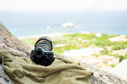 (Selective focus) Professional camera with a super telephoto lens with camouflage cover on a rocky mountains. Paparazzi equipment, Costa Smeralda, Sardinia, Italy.