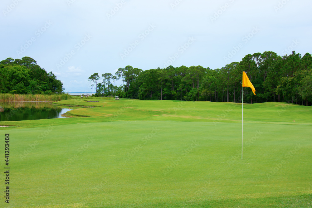 Obraz premium Lush golf course green with yellow flag in the hole and fairway with water trap, sand traps and tree line in central Florida on a crisp summer morning.
