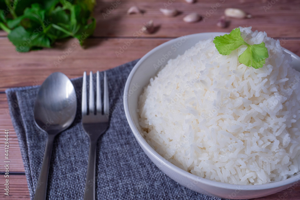Steam organic white rice in a white ceramic bowl with silver cutlery and apron and green coriander stained on a brown wooden table. Steam organic white rice in a white ceramic bowl with silver cutlery and apron and green coriander stained on a brown wooden table.