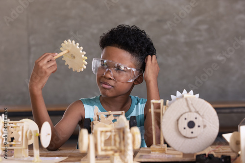 Happiness cheerful child curly black hair clever wearing safety glasses looking parth of  working model of a mechanism made of wood on table.Education and innovation concept. photo