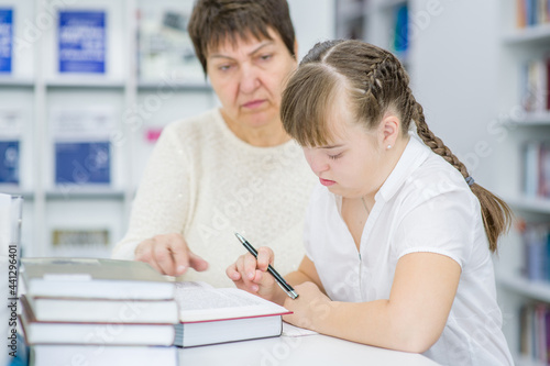 Old woman helps to girl with syndrome doing homework at school. Education for disabled children concept © Ermolaev Alexandr