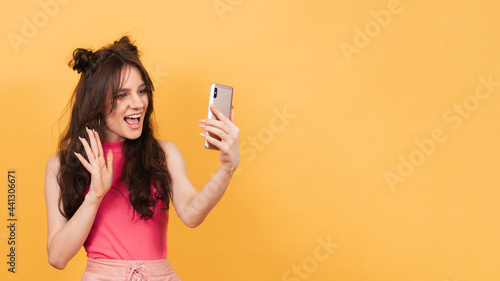 A smiling woman holds a smartphone on which she records a video message or talks in a video chat, standing on a yellow background. A copy of the space. Space for text