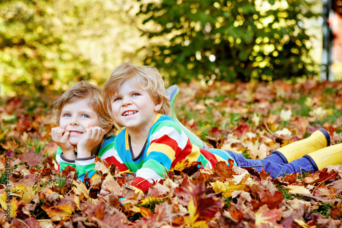 Two little twin kids boys lying in autumn leaves in colorful fashion clothing. Happy siblings having fun in autumn park on warm day. Healthy children with blond hairs and blue eyes with maple foliage.