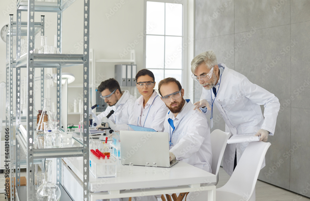 Senior male supervisor and young scientist assistant group discussing experiment test result looking at laptop screen. Researchers team developing new drugs working at pharmacy laboratory Senior male supervisor and young scientist assistant group discussing experiment test result looking at laptop screen. Researchers team developing new drugs working at pharmacy laboratory