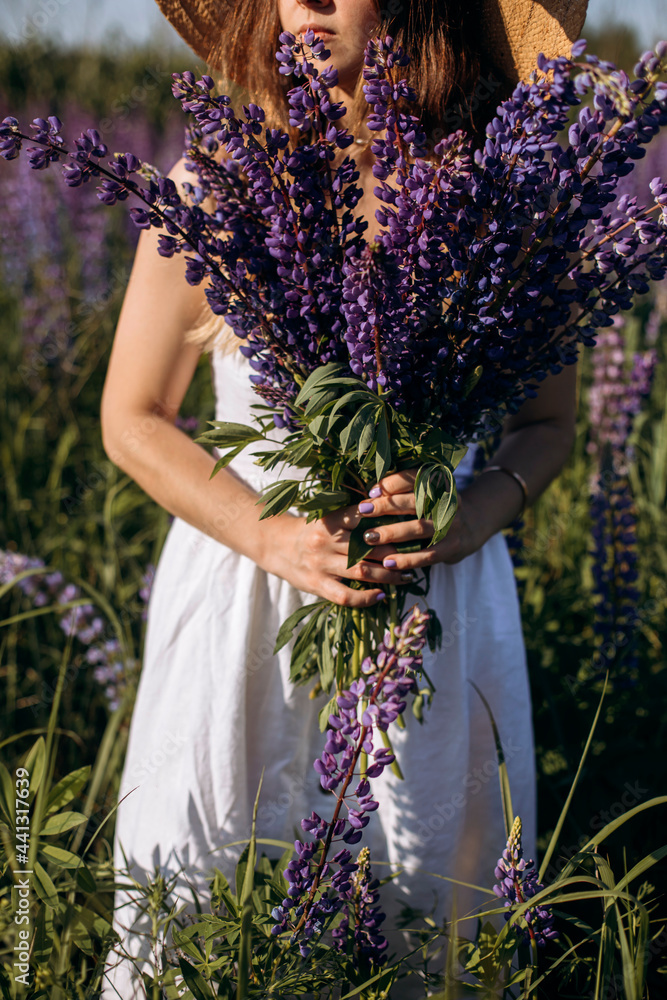 a young woman in a straw hat holds a bouquet of wild flowers in her hands. a girl walks through a lupine field on a bright sunny day. girl in a white dress in a lavender field.